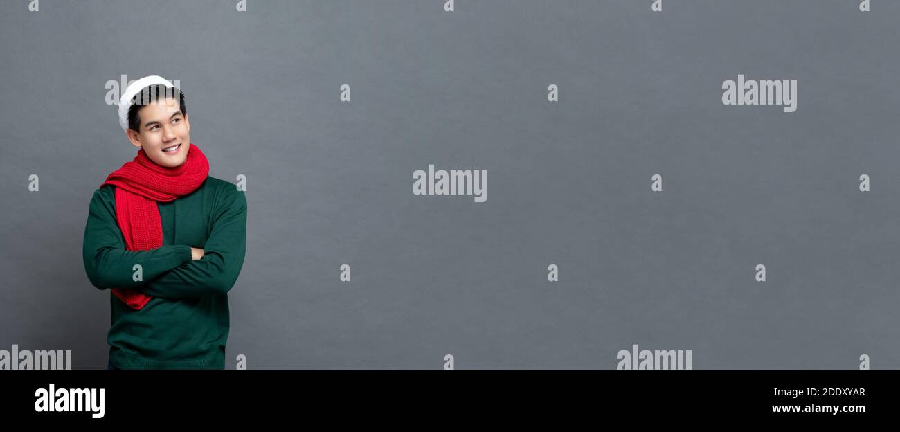 Giovane bell'uomo asiatico in felpa di Natale verde e rosso sciarpa sorridente e guardando lo spazio di copia con le braccia barrato isolato sul dorso grigio dello striscione Foto Stock