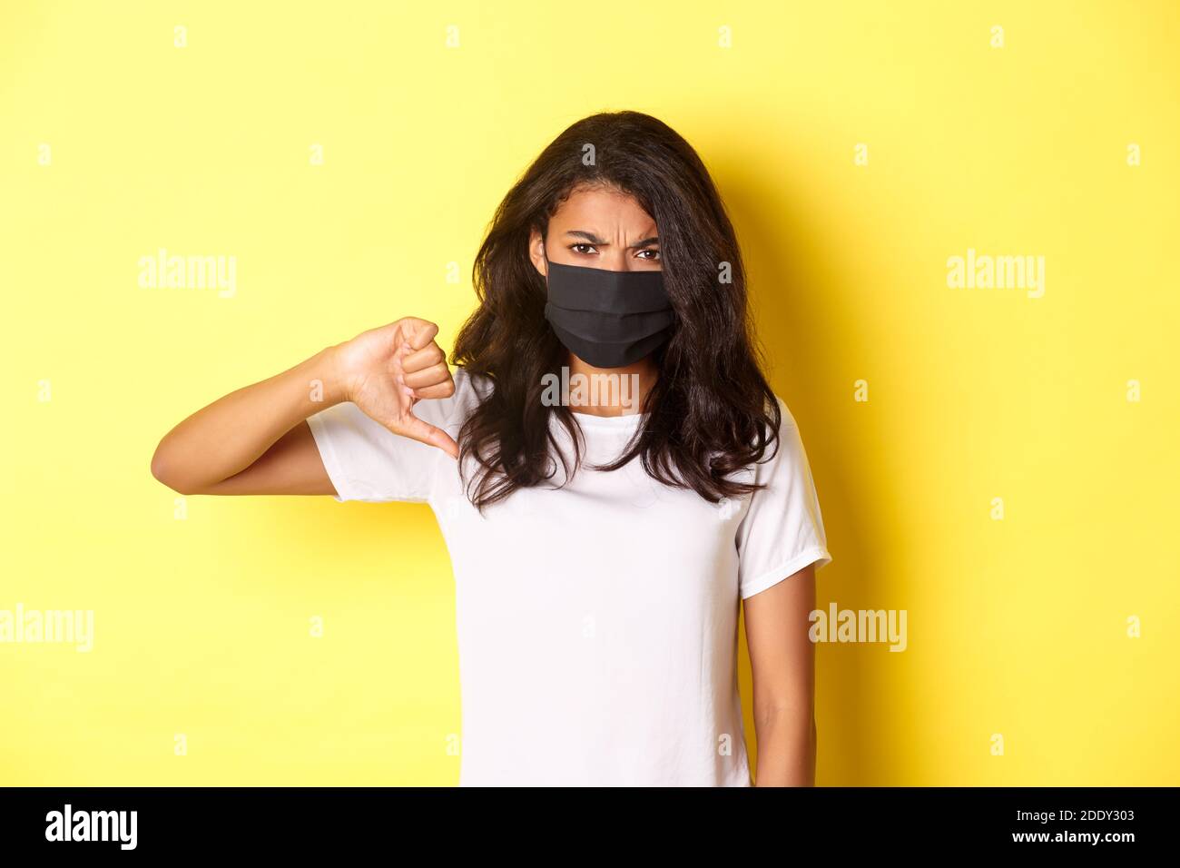 Concetto di covid-19, divaricamento sociale e stile di vita. Immagine della delusa modello femminile afro-americana, indossando maschera nera, mostrando pollici Foto Stock