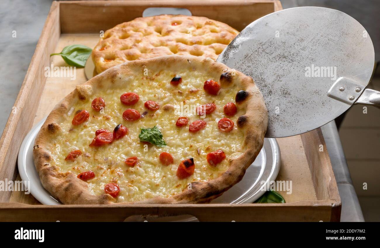 Pizza italiana con pomodori ciliegini e mozzarella, particolare della pala da forno che scarica la pizza appena sfornata su vassoio di legno Foto Stock