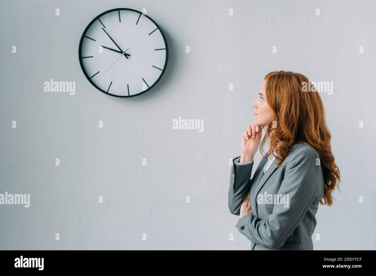 Vista laterale di una donna d'affari premurosa che guarda l'orologio a parete grigio Foto Stock