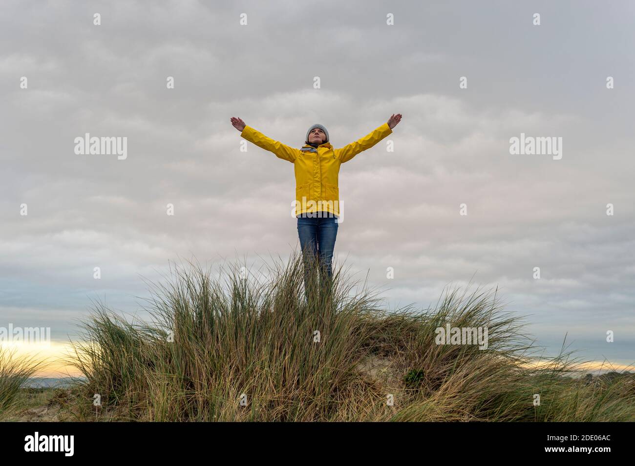 Donna che indossa un cappotto giallo sulla cima di una duna di sabbia con le braccia sollevate, concetto di successo. Foto Stock
