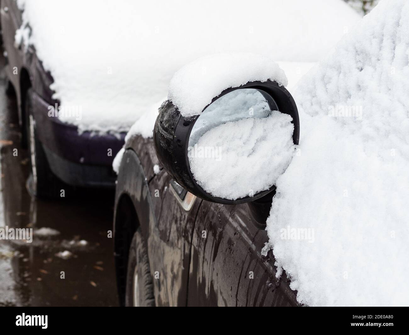 la prima neve sullo specchio retrovisore laterale della vettura nera parcheggiata lungo la strada della città in freddo giorno d'autunno Foto Stock