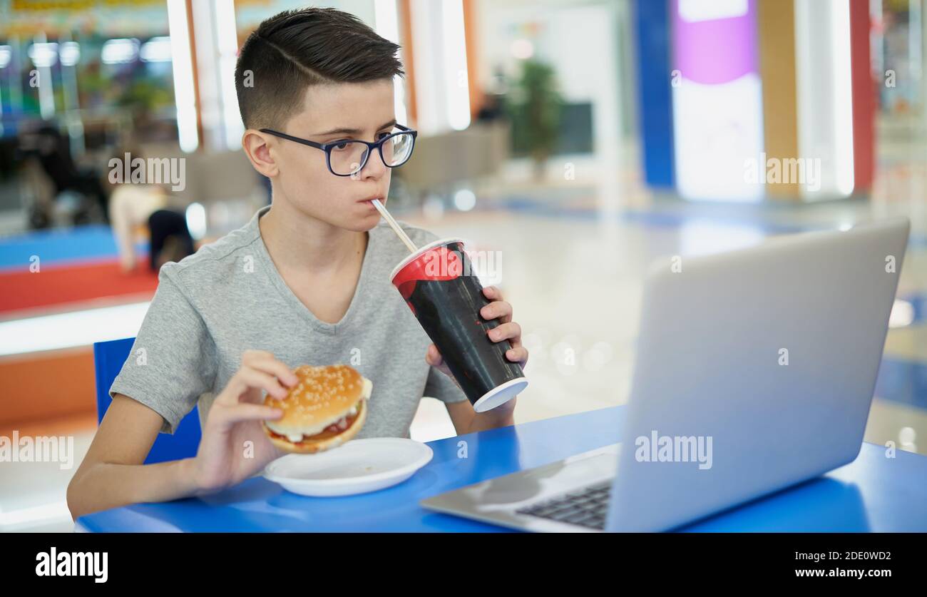 Adolescente ragazzo mangiare malsano fast food e bere soda mentre seduto in un'area ristoro Foto Stock