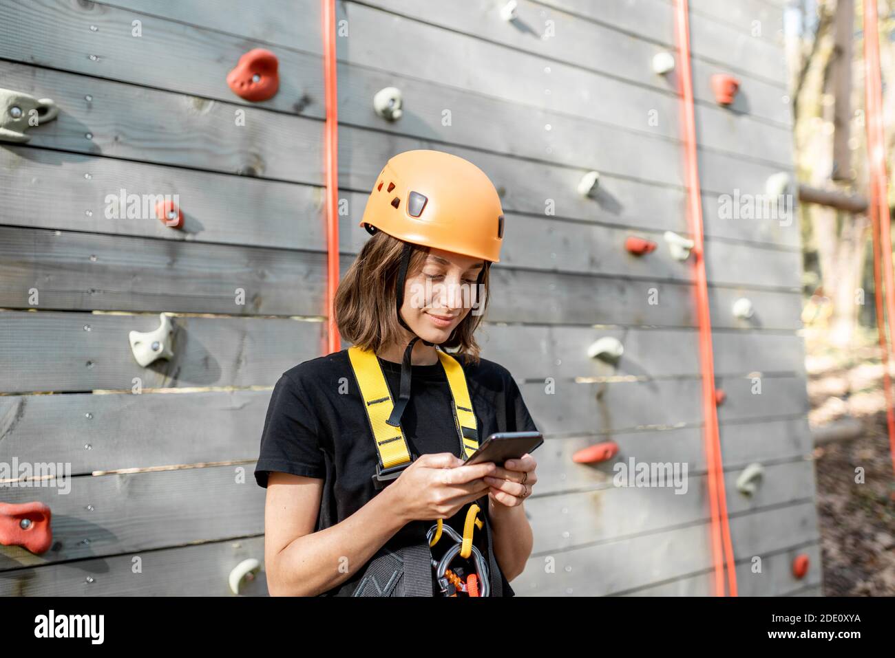 Giovane donna in attrezzature sportive di protezione con uno smartphone vicino alla parete di arrampicata all'aperto Foto Stock