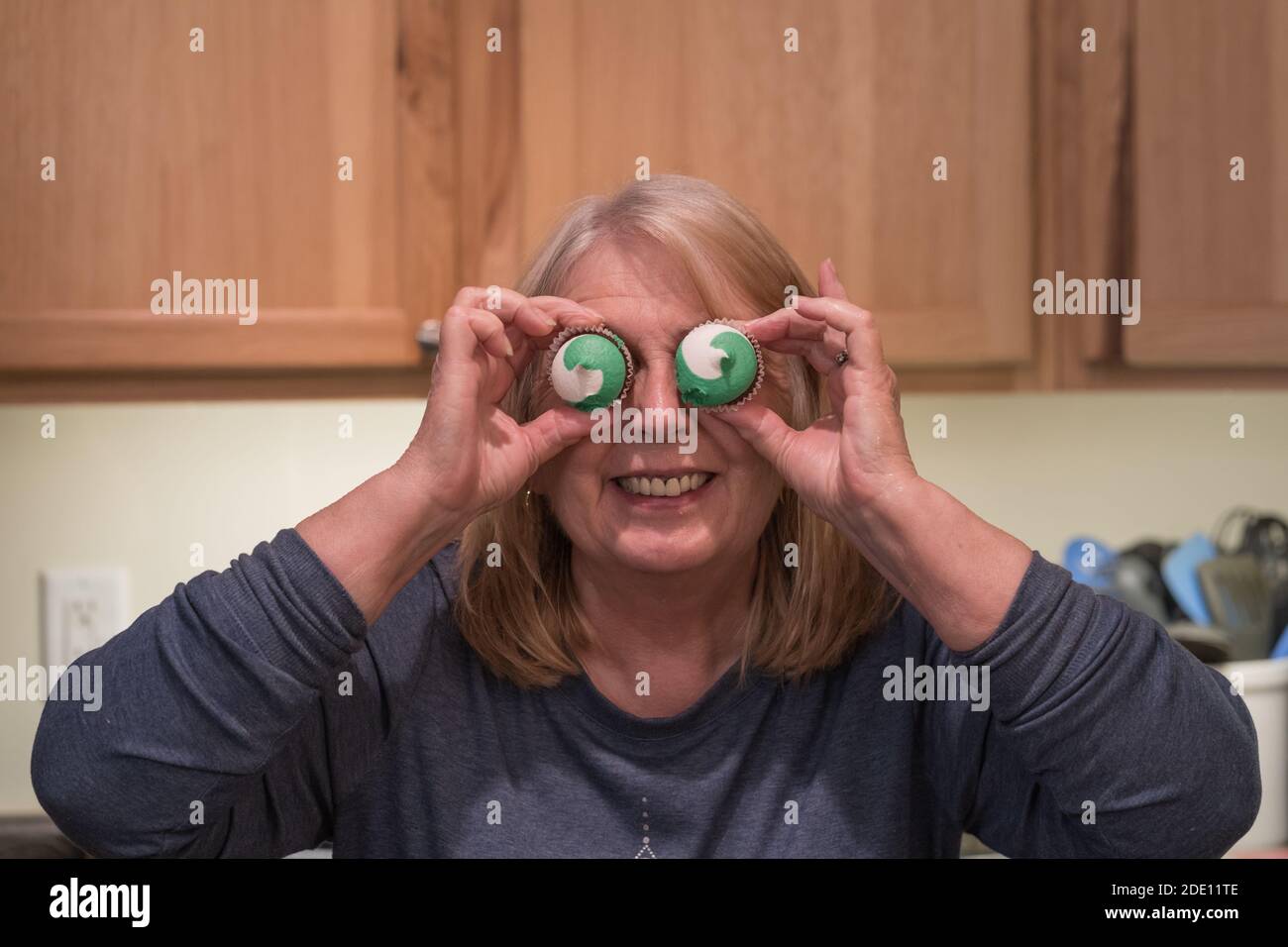 Baby boomer donna tiene due mini cupcakes ghiacciati fino ai suoi occhi, come occhiali, comportandosi sciocco Foto Stock