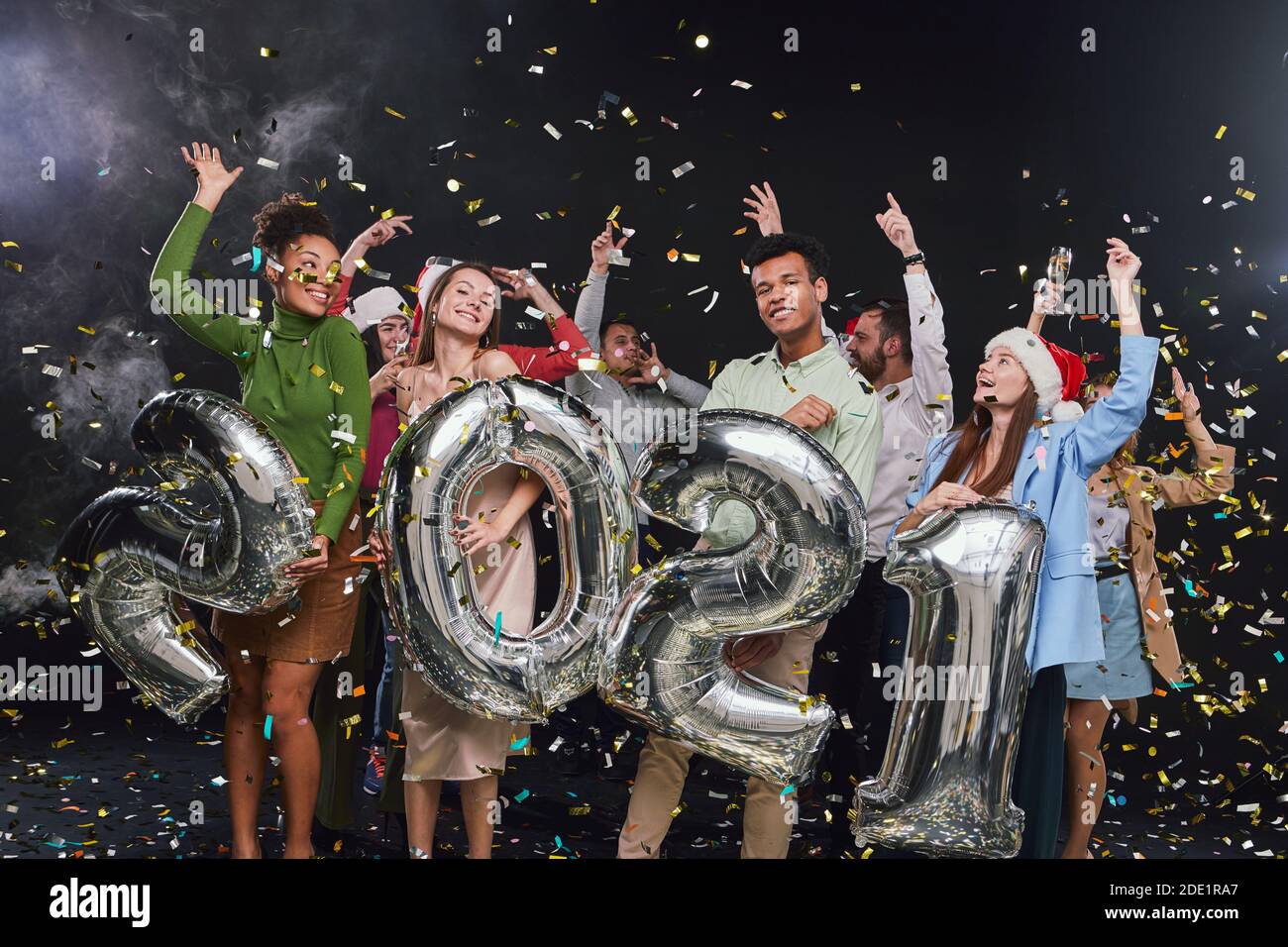 Gruppo di giovani felici multirazziali che reggono palloncini d'argento in forma di numeri 2021 e ballano, i confetti che cadono nell'aria, celebrando la vigilia di Capodanno con gli amici. Festa di natale Foto Stock