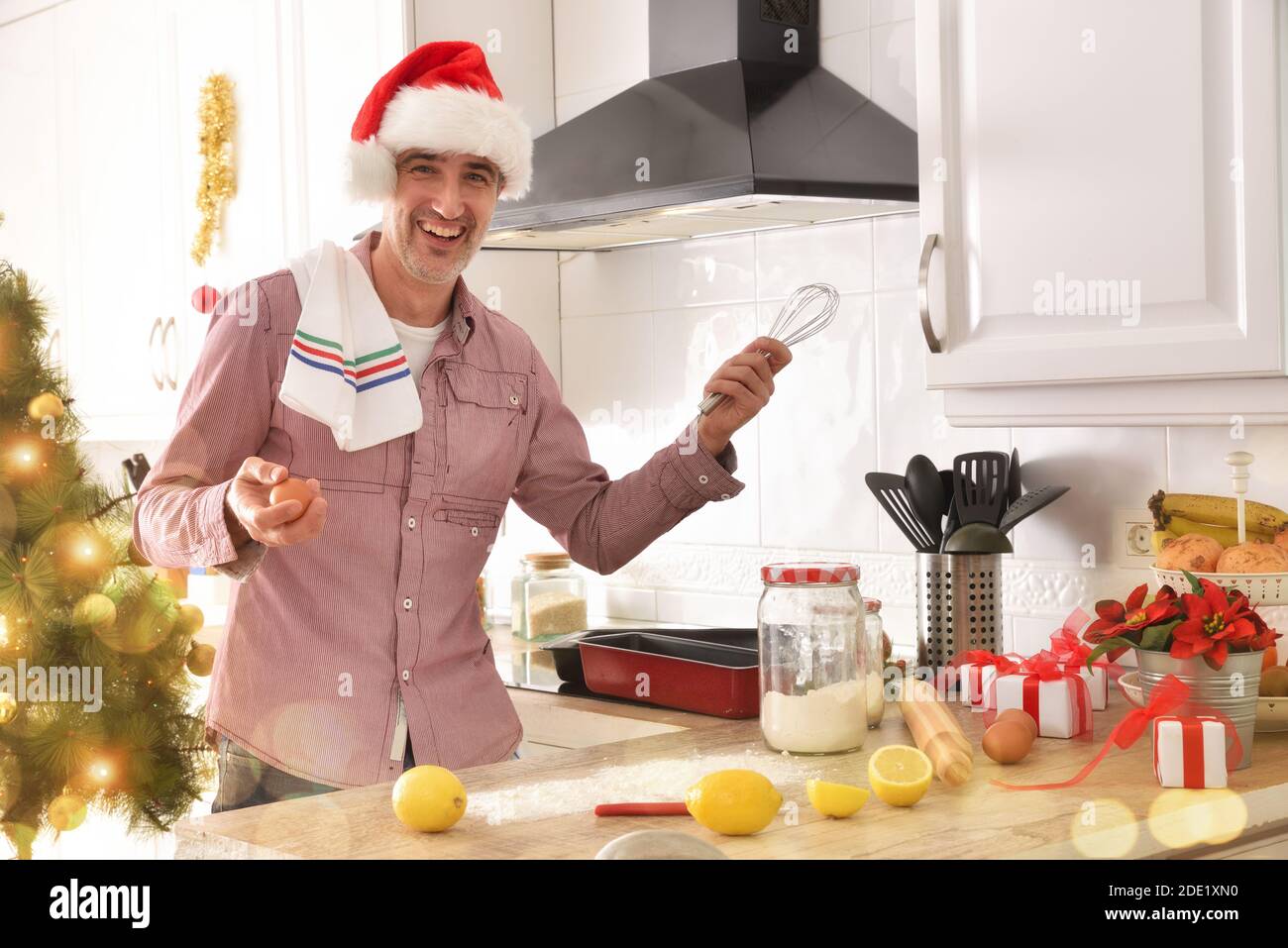 Uomo ridente preparato a cucinare un dessert con gli ingredienti sopra la panca della cucina con cappello di natale e sfondo di albero con luci Foto Stock