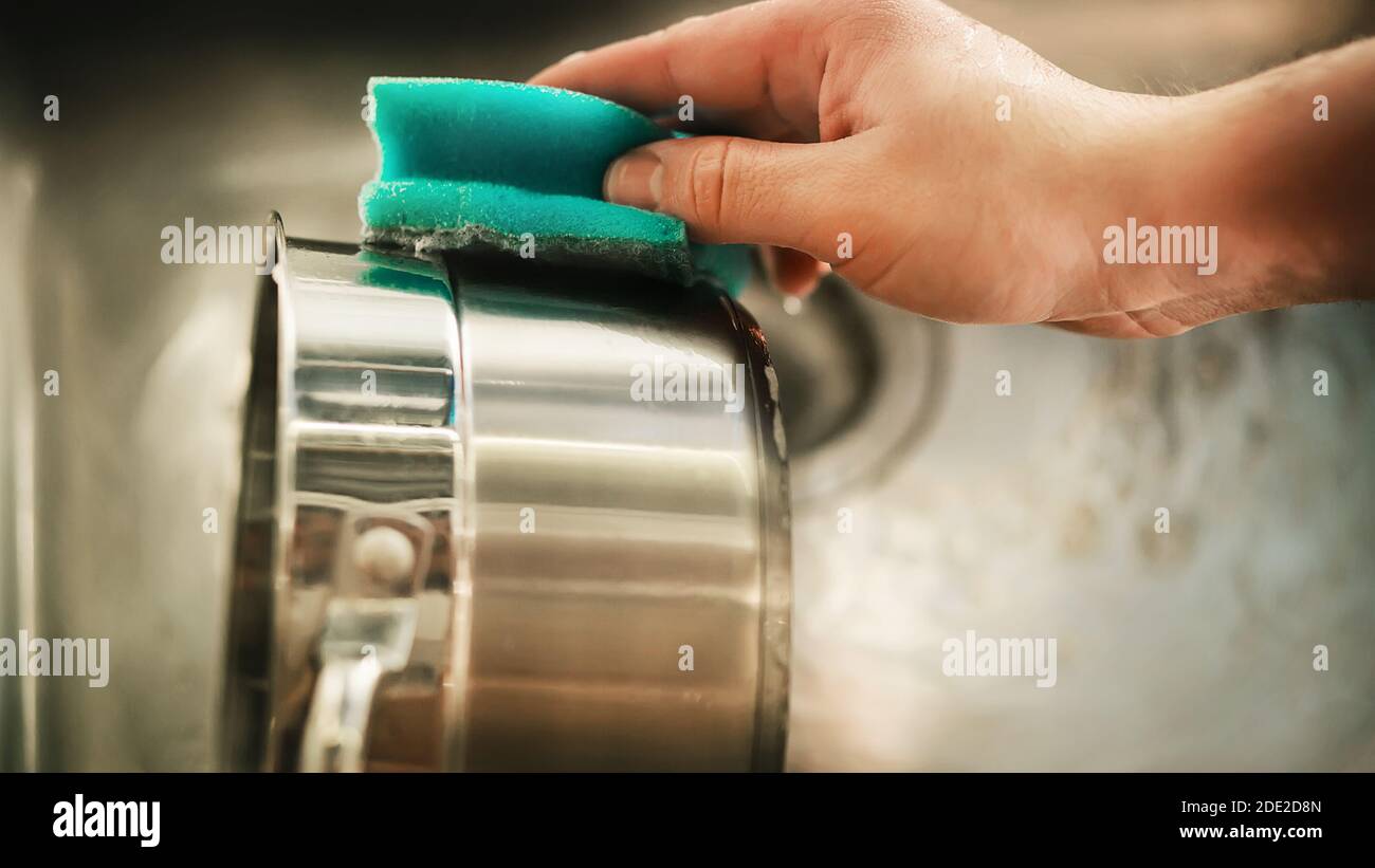 Un uomo tiene in mano una spugna blu saponata e la lava per lavare una padella metallica sopra il lavandino della cucina. Le faccende domestiche. Utensili da cucina. Foto Stock