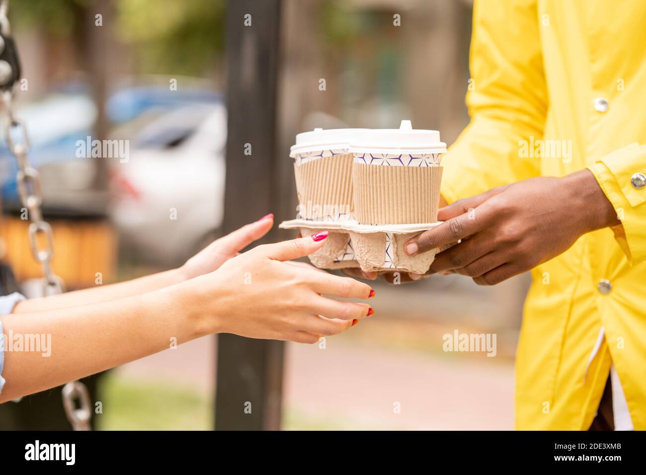 Mani di corriere africano maschio in uniforme gialla che passa due bicchieri di caffè dal ristorante o caffè alla giovane donna in ambiente urbano Foto Stock