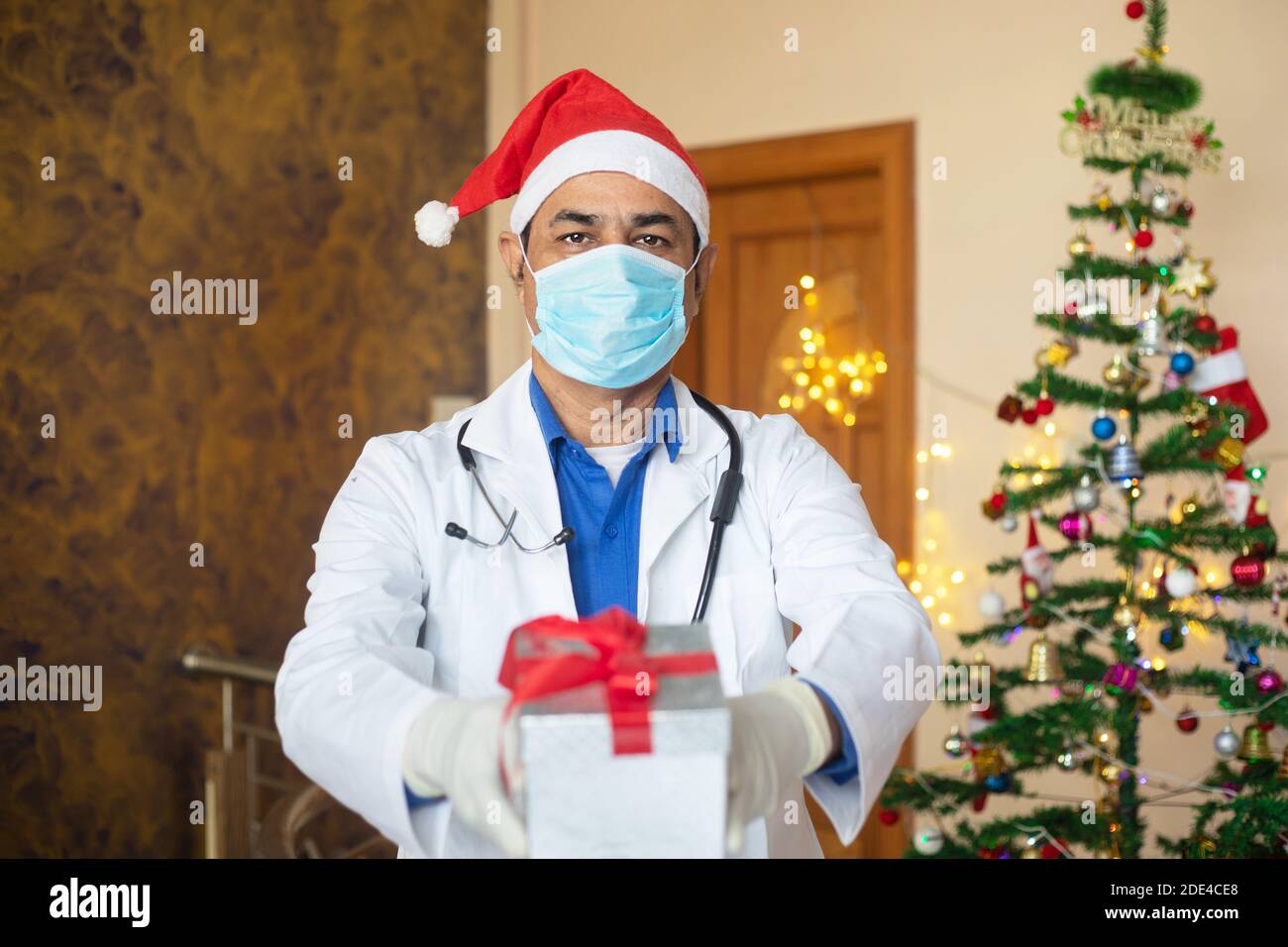 Medico indossando maschera e cappello di santa mostrando confezione regalo di natale, celebrazione durante la pandemia covid-19, nuovo stile di vita normale. Sanità e medico. Foto Stock