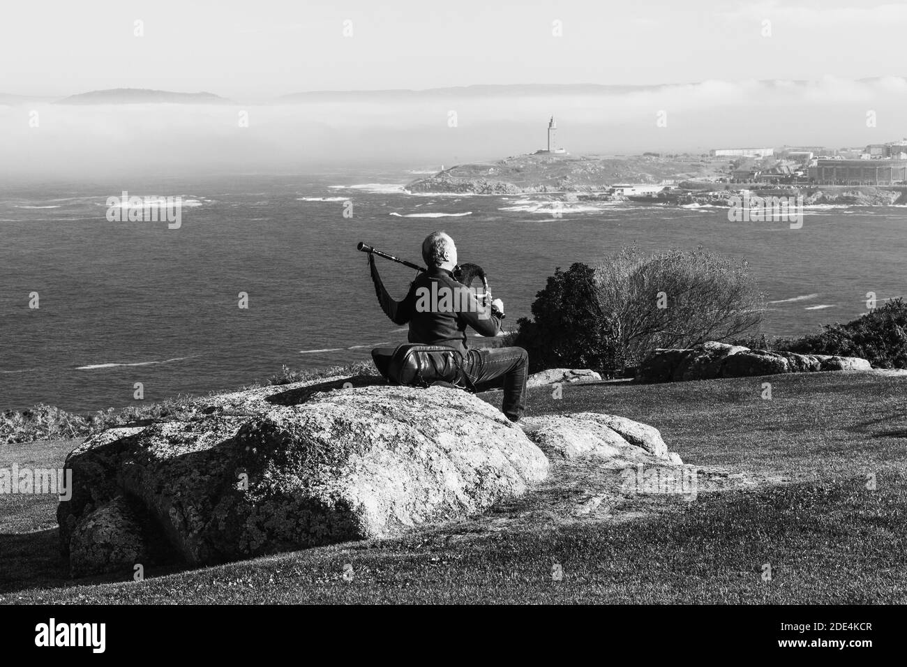 vista da dietro del bagpiper seduto su un gioco di rock Le cornamuse con la Torre di Ecole sullo sfondo tra la nebbia Foto Stock