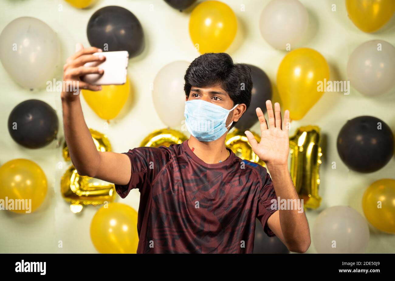 Giovane uomo con maschera medica occupato facendo videochiamata su mobile oltre 2021 nuovo anno decorati sfondo - concetto di quarantena festeggiamenti di vacanza dovuto Foto Stock