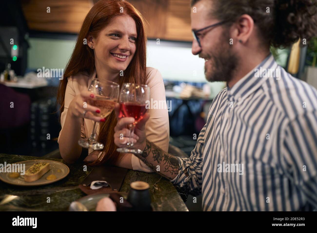 giovane uomo caucasico tatuato con una barba bere vino con una bella femmina rossa a cena romantica in ristorante, tostare, guardarsi l'un l'altro Foto Stock