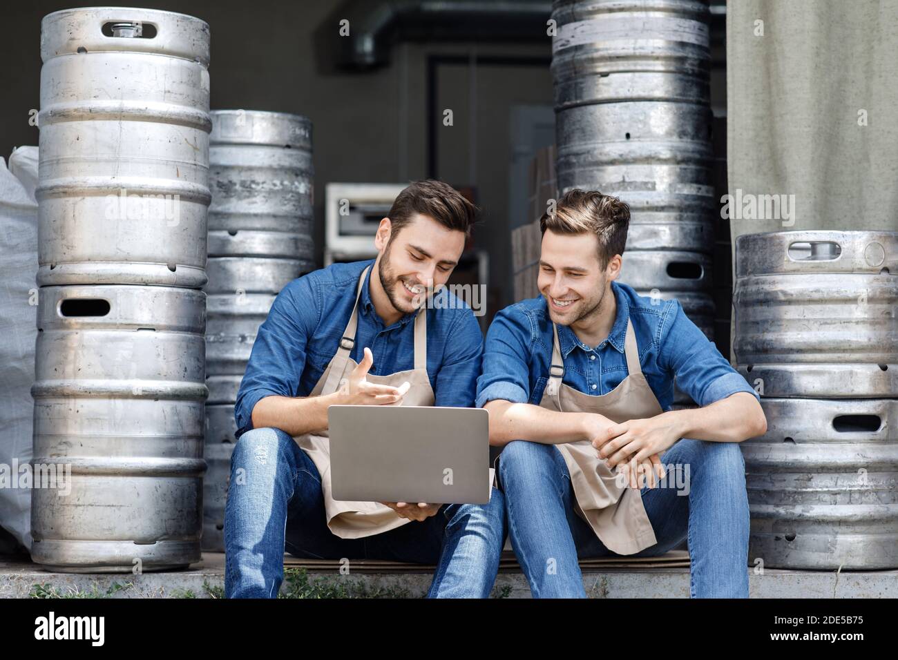 Felici ragazzi millenari, proprietari in grembiuli siedono sul pavimento, guardare i dati in un notebook e parlare Foto Stock