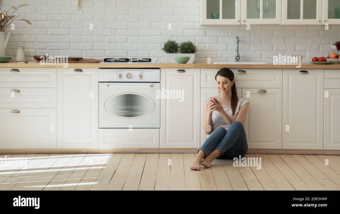 Giovane donna sorridente seduta sul moderno pavimento della cucina, utilizzando uno smartphone Foto Stock