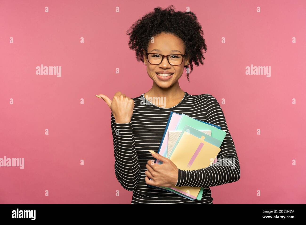 Tutor afro-americano o insegnante donna isolata su parete rosa studio. Studentessa indossa occhiali che tengono notebook, sorridendo, mostrando il dito sullo spazio libero. Foto Stock
