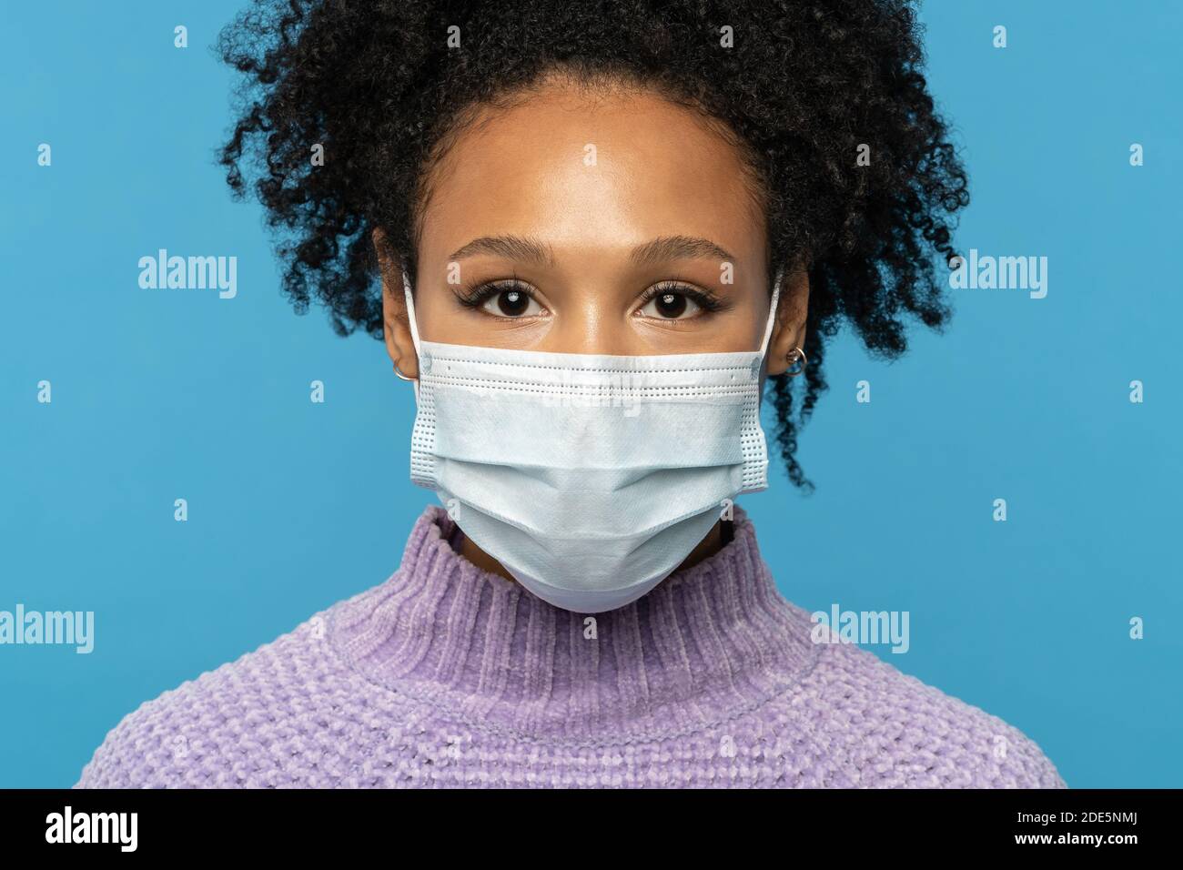 Primo piano ritratto studio di donna afroamericana indossare maschera medica viso durante covid-19, epidemia di coronavirus o stagione influenzale, isolato su sfondo blu Foto Stock