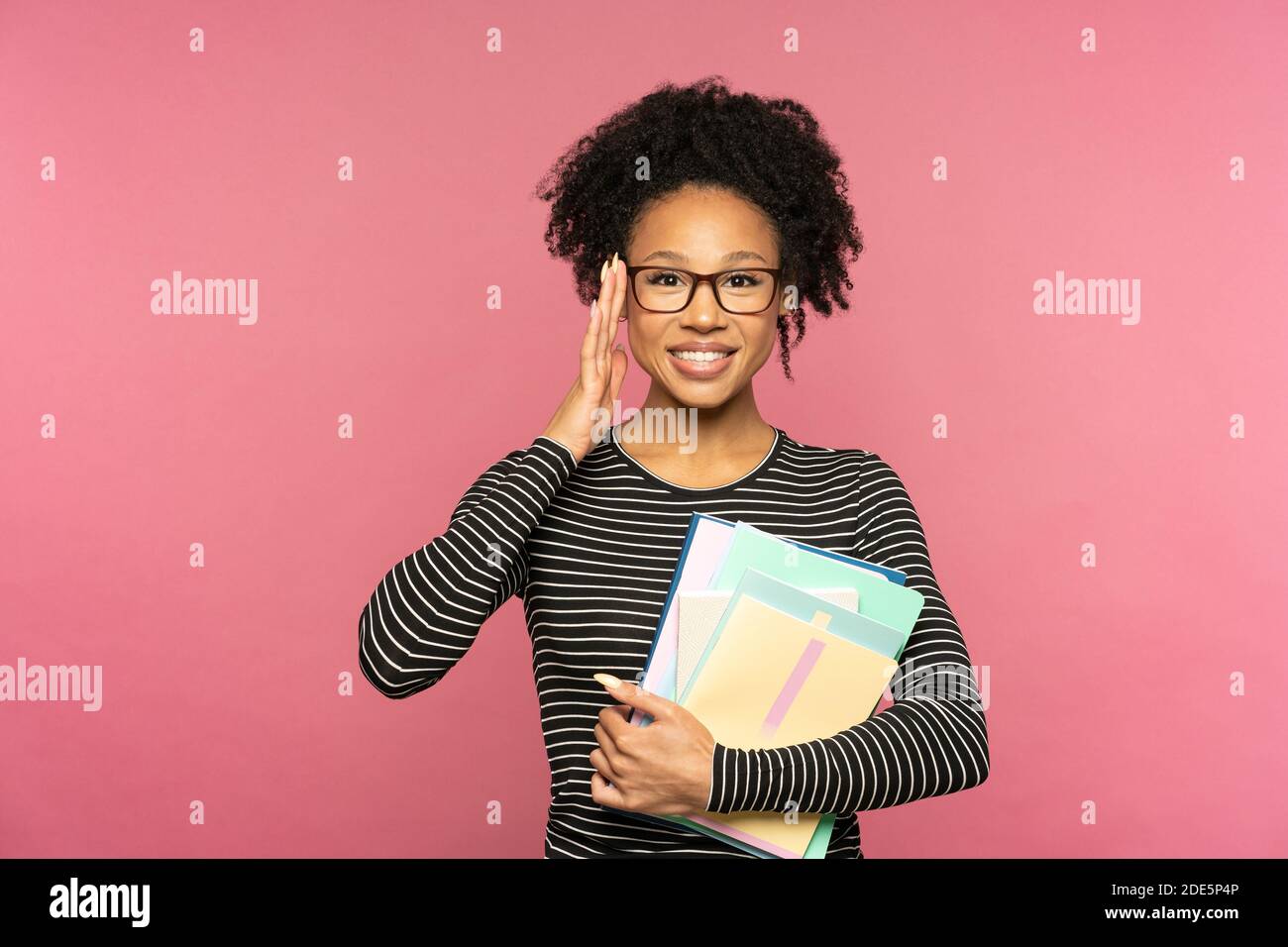 Giovane felice tutor afro-americano o insegnante donna isolata su muro rosa studio. Studentessa indossa occhiali che tengono notebook e sorridono. Istruzione in h Foto Stock
