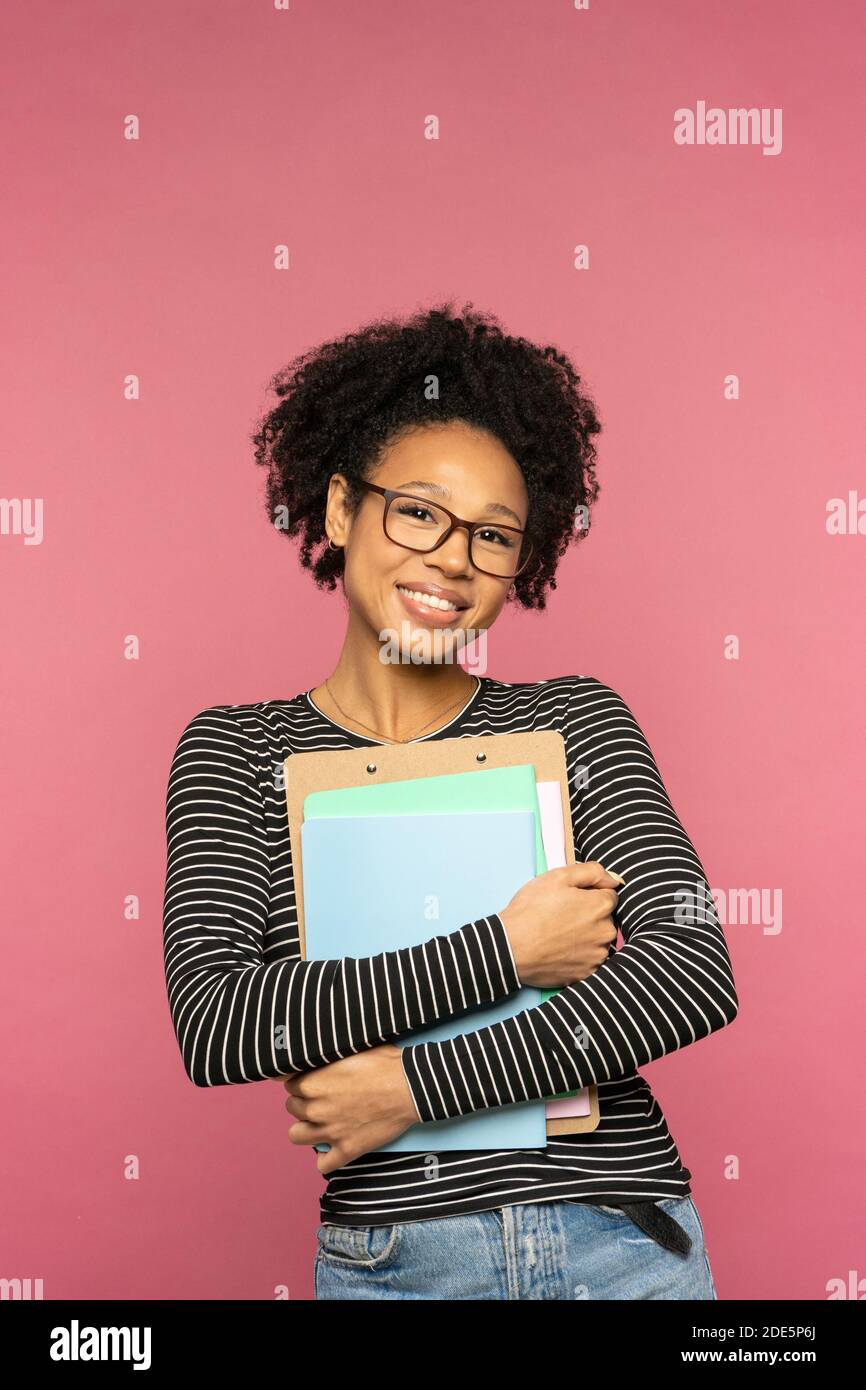 Giovane felice tutor afro-americano o insegnante donna isolata su muro rosa studio. Studentessa indossa occhiali che tengono notebook e sorridono. College e Ciao Foto Stock