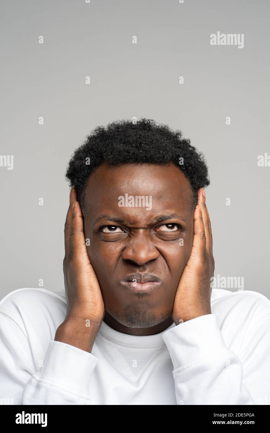 Closeup arrabbiato pazzo stressato uomo afro che copre le orecchie e guardando in su, sente fastidioso rumore forte da vicini al piano superiore isolato su sfondo grigio studio Foto Stock