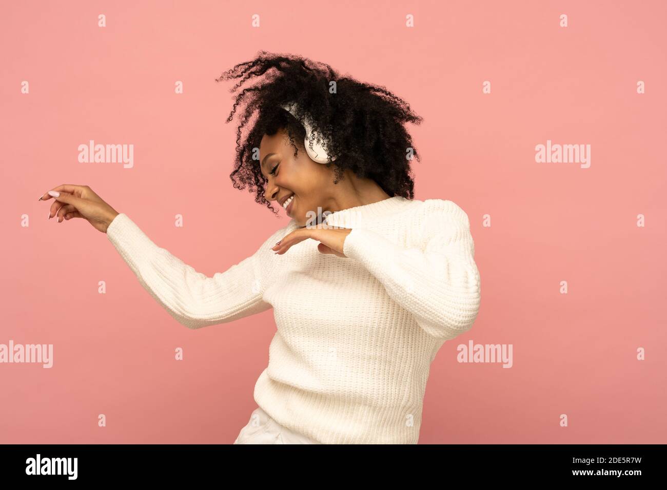 Donna biraciale energizzata in pullover bianco che canta, indossa cuffie wireless, ha un buon umore, isolato su sfondo rosa. Felice pelle scura riccia Foto Stock