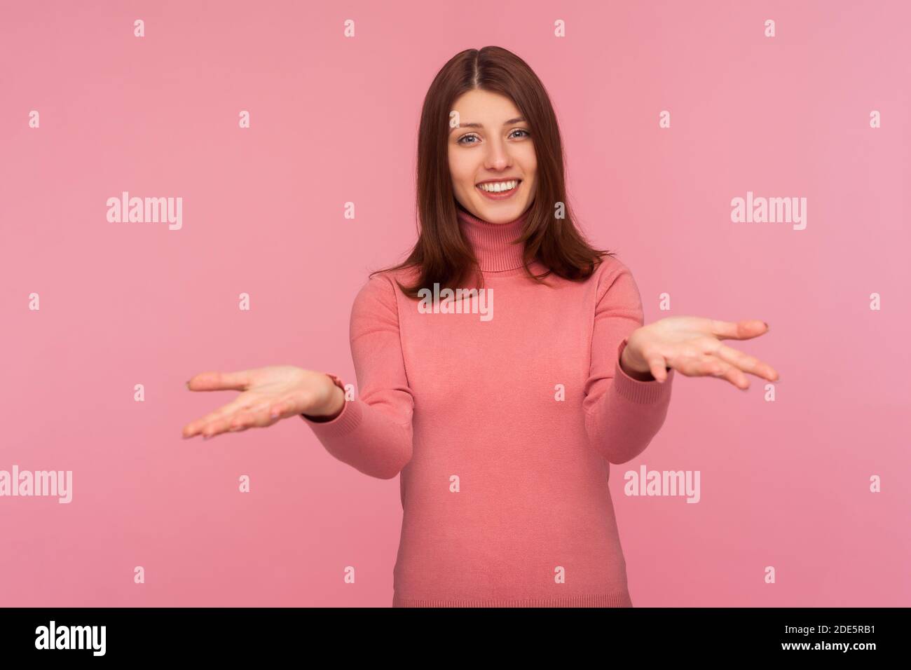 Donna accogliente a cuore aperto con capelli marroni in maglia rosa in piedi con mani vuote aperte, felice di condividere, dando tutto ciò che ha. Studio interno sh Foto Stock