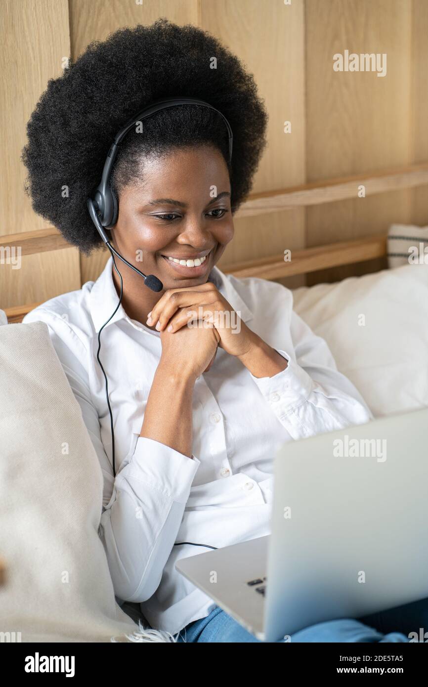 Sorridente donna afro-americana millenaria che indossa cuffie, comunica tramite chat video sul laptop, parla in zoom, guarda webinar o video streaming conf Foto Stock