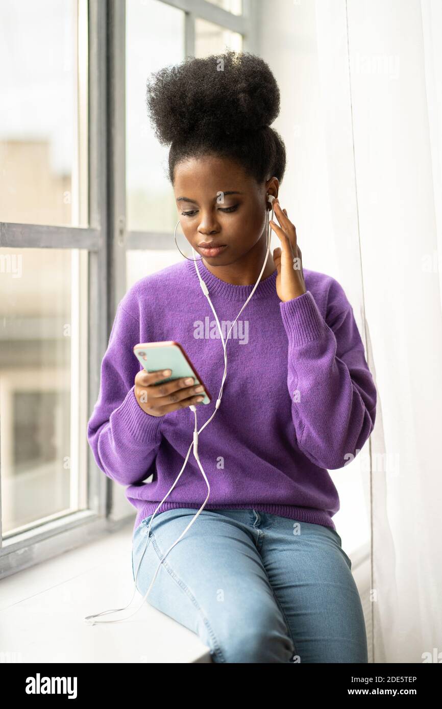 Giovane donna millenaria afroamericana con maglione di abbigliamento afro-acconciato, seduta sul davanzale, ascolta la musica con le cuffie cablate sul cellulare smar Foto Stock