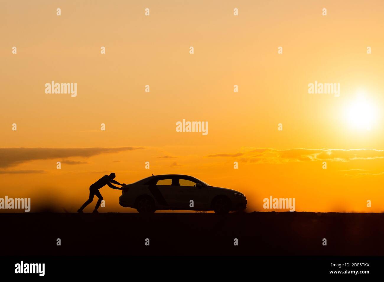 Problemi di trasporto. Silhouette di uomo driver che spinge la sua auto lungo una strada vuota dopo il guasto al tramonto, spazio di copia, vista laterale. Foto Stock