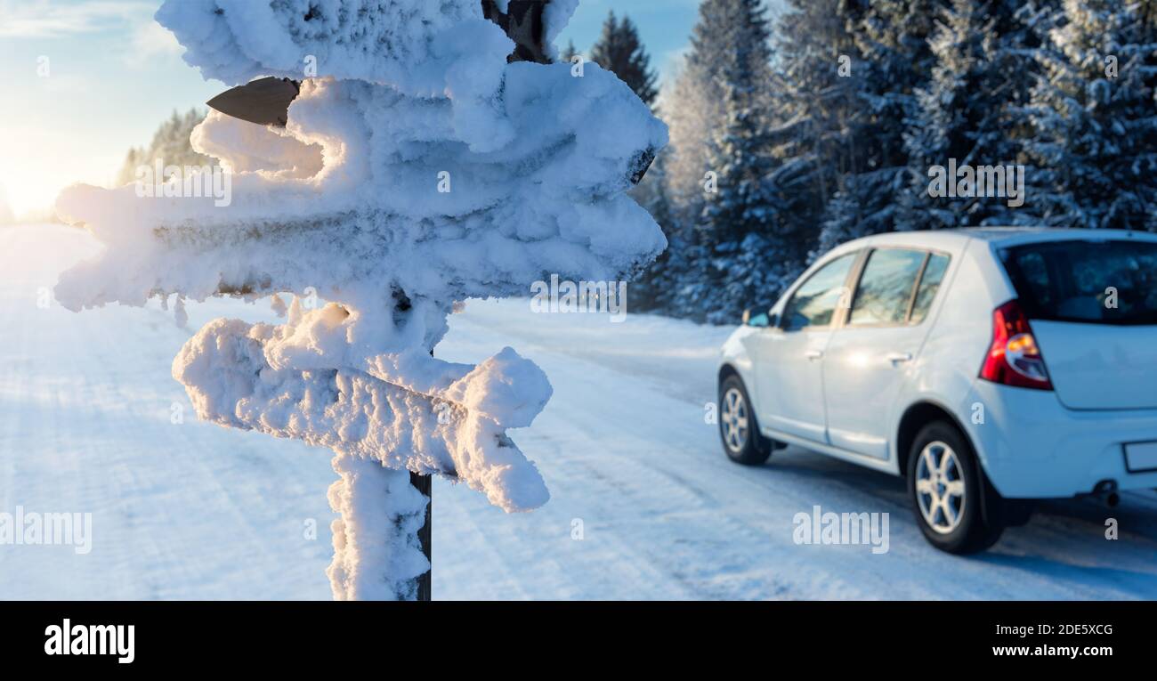 Cartello coperto di neve con frecce in legno sullo sfondo di una macchina su una strada. Viaggio invernale Foto Stock