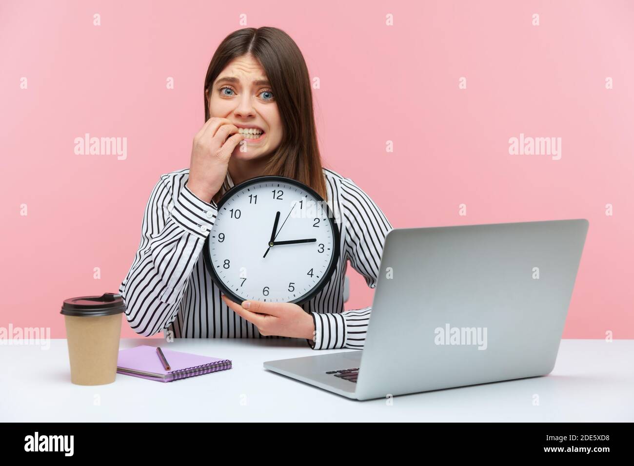Impaziente donna nervosa in camicia a righe seduta sul posto di lavoro mordente le sue unghie tenendo grande orologio da parete, scadenza. Studio al coperto isolato su b rosa Foto Stock