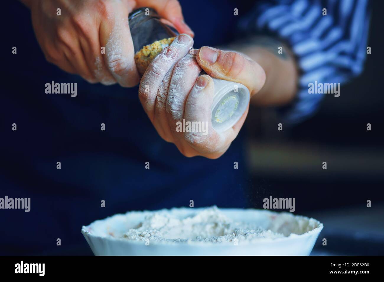 Uno chef in grembiule blu e camicia a righe tiene un bicchiere di sale shaker con condimento e lo versa in una ciotola di farina per la futura cottura. Cucina casalinga. Foto Stock