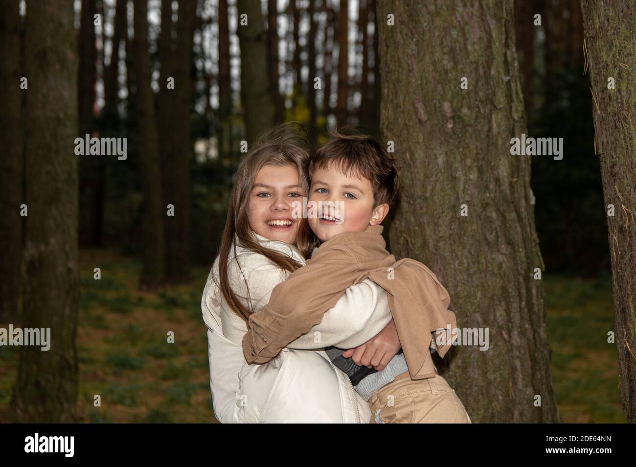 Una ragazza adolescente in un vestito leggero abbraccia il suo fratello minore. I bambini sono felici e ridono tra gli alberi della foresta. Vera infanzia e famiglia happine Foto Stock