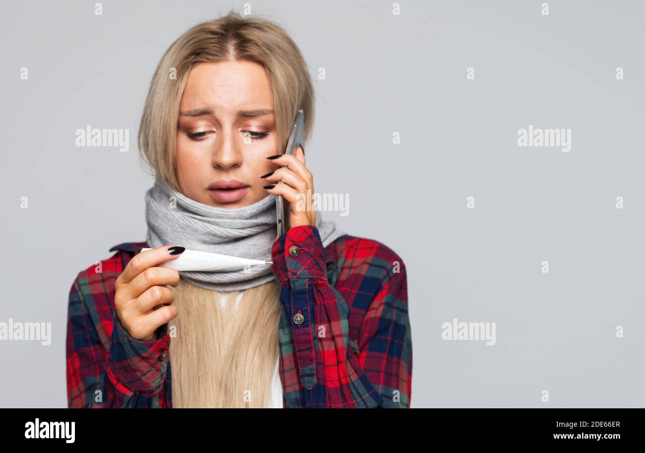 Malata giovane donna che guarda il termometro, parlando al telefono, ha sintomo di influenza, isolato su sfondo grigio studio. Foto Stock