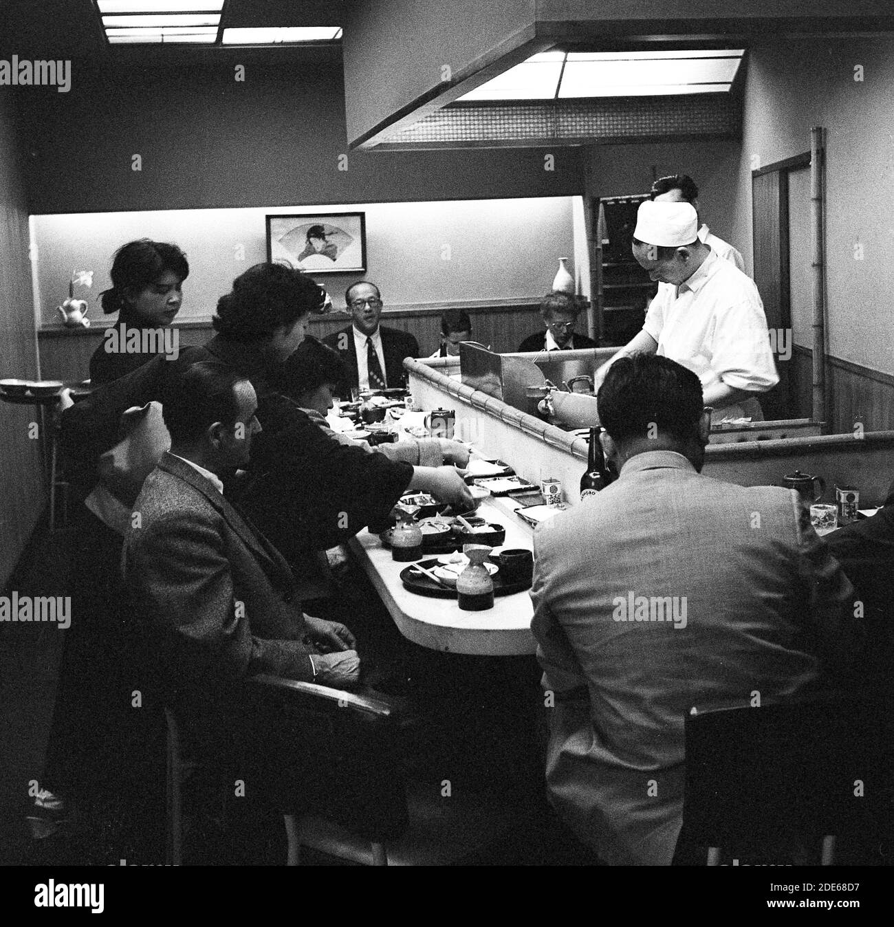 Anni '1960, storico, all'interno di un piccolo sushi bar, persone sedute su sgabelli da bar in un bancone per guardare lo chef di fronte a loro preparare il cibo, Tokyo, Giappone. Foto Stock