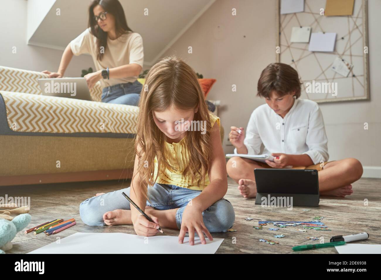 Bambina seduta sul pavimento e disegnando mentre suo fratello fa i compiti in linea su tablet digitale, giovane bella madre seduta sul divano con Foto Stock