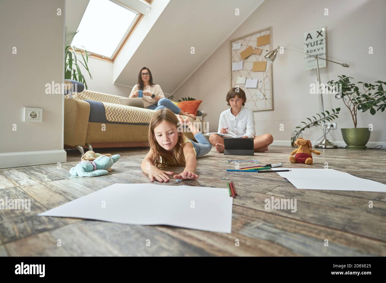 Prescolare ragazza sdraiata sul pavimento e disegnare con matite colorate, mentre suo fratello fare i compiti online, madre seduta su divano con tazza di caffè Foto Stock