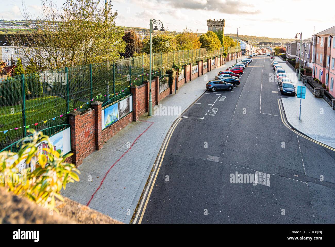 Pannelli fotografici storici allineano un muro di mattoni lungo una strada in pendenza nella zona di Bogside di Londonderry, contea di Derry, Irlanda del Nord; una torre delle antiche mura cittadine si erge sullo sfondo. Le auto sono parcheggiate lungo la carreggiata. Foto Stock