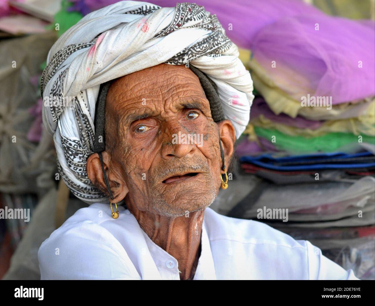 Il vecchio Rabari indiano senza denti fragile con orecchini tribali dorati e il viso rugoso indossa un turbante tradizionale e guarda lo spettatore. Foto Stock