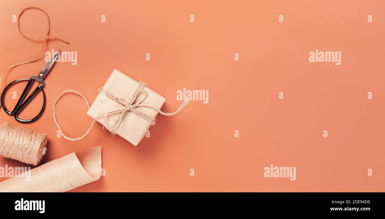 Confezione regalo in carta artigianale su sfondo rosso caldo, forbici e carta da imballaggio nelle vicinanze. Il concetto di preparare i regali per la festa. Vista dall'alto, spazio di copia Foto Stock
