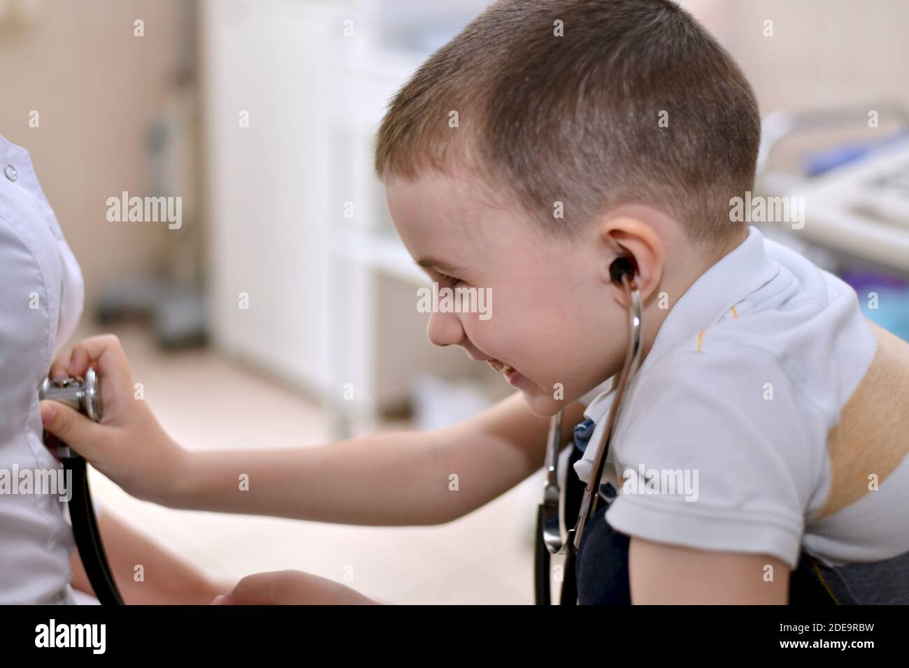 Il bambino ride in imbarazzo con le cuffie stetoscopio nelle orecchie, e applica la membrana audio testa al cappotto bianco. Comunicazione divertente Foto Stock