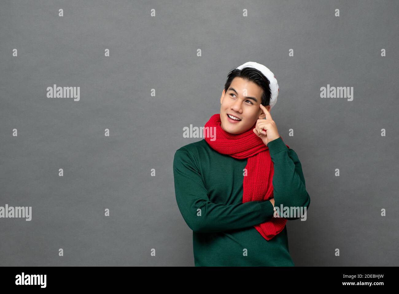 Curioso bell'uomo asiatico che indossa abiti a tema natalizio guardando da parte lo spazio vuoto e pensando con la mano sulla testa, studio girato isolato su grigio Foto Stock