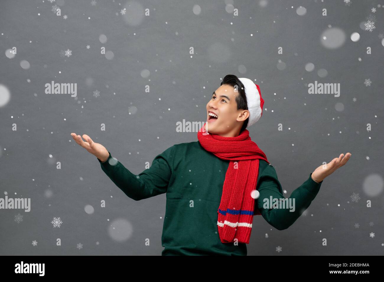 Giovane uomo asiatico divertito che indossa abiti a tema natalizio con aperto gesto delle palme in nevicata Foto Stock