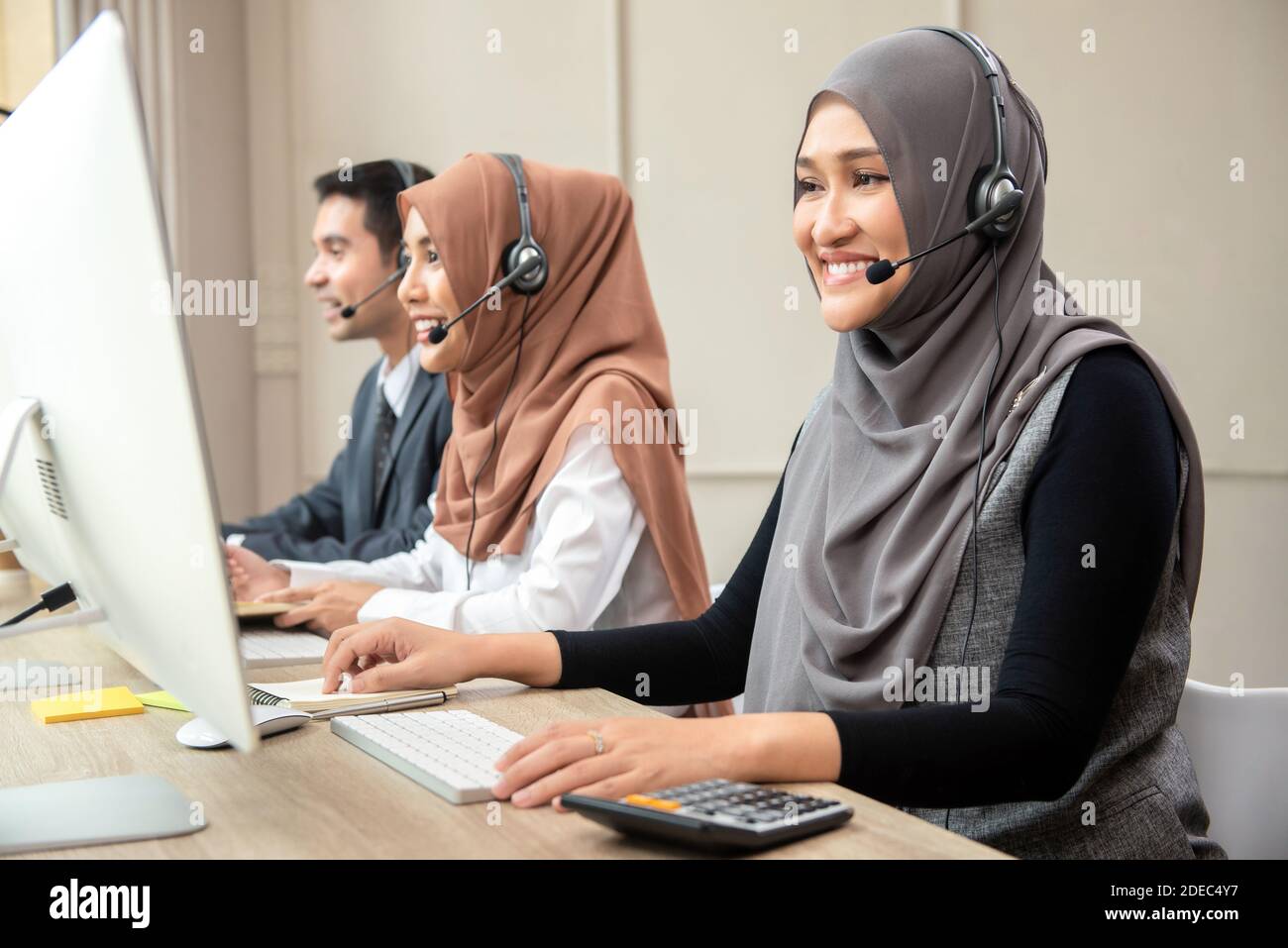 Donna musulmana asiatica sorridente che indossa cuffie con microfono che lavorano come cliente operatore sanitario con team in ufficio del call center Foto Stock