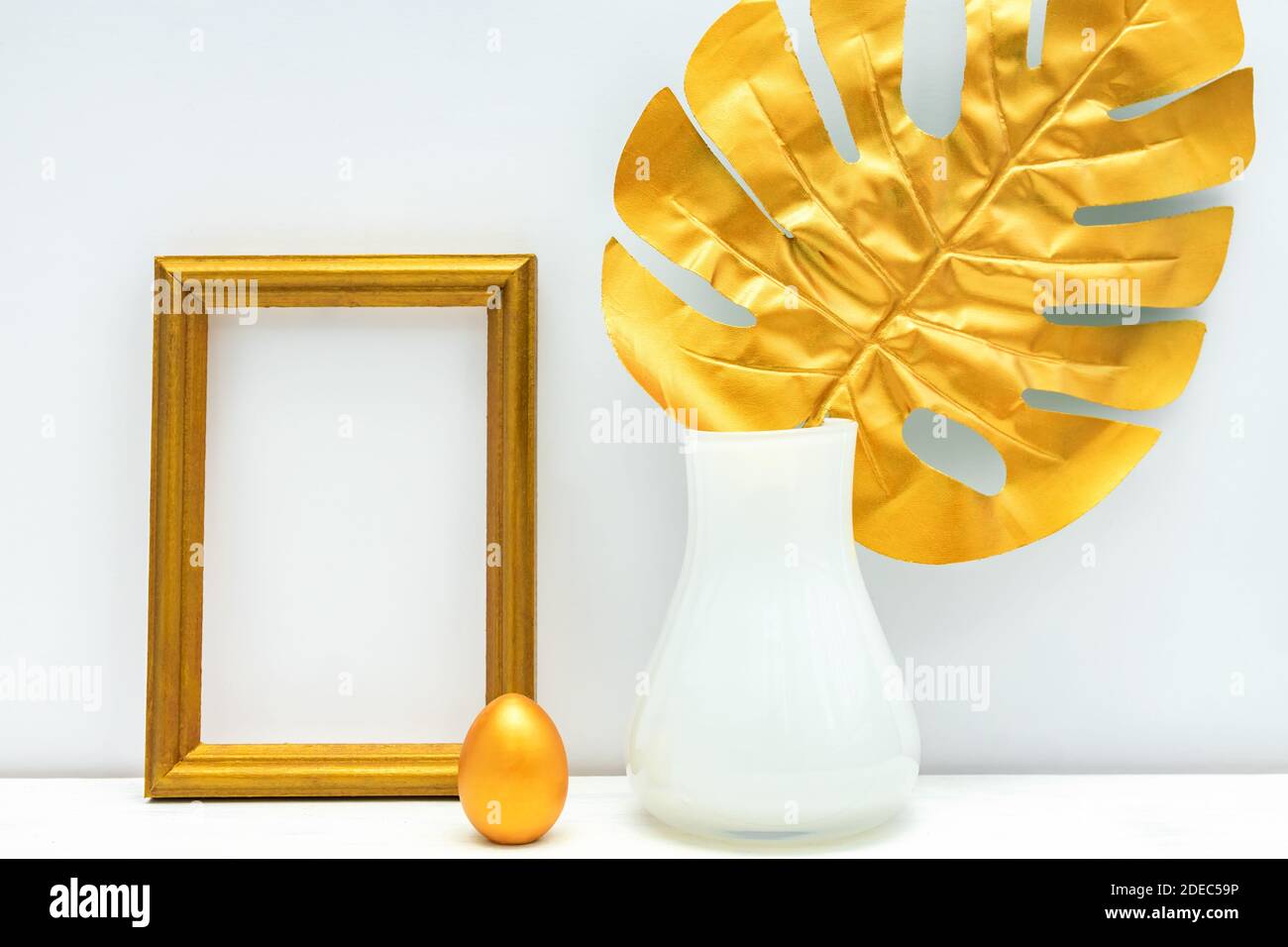 design mockup interno in oro e bianco. photoframe vuoto, uovo verniciato e foglia di monstera in vaso bianco su sfondo bianco della parete. Design elegante e lussuoso. Foto Stock