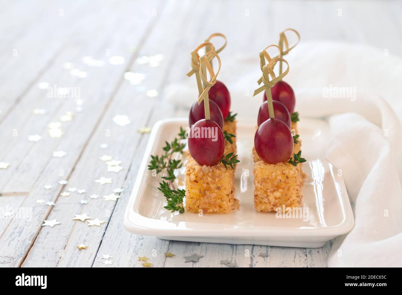 Canape con uva rosa e formaggio in noce impanata, su spiedini di legno. Spuntino festivo su un piatto bianco su sfondo di legno azzurro, selettivo Foto Stock