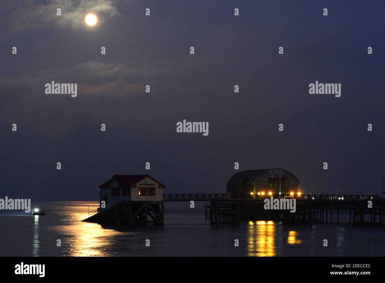 Luna piena che si riflette sul mare a Mumbles. Il molo, la stazione di Lifeboat e una barca da pesca sono illuminati dal chiaro di luna e ultimo raggi del sole che tramonta. Foto Stock