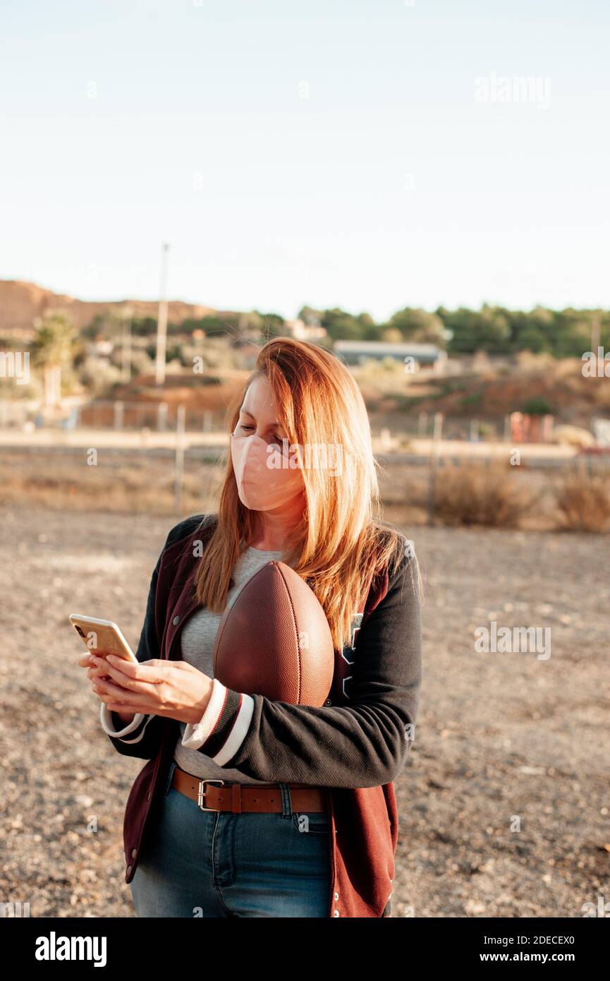 Donna che indossa una maschera protettiva per il viso e usa il suo smartphone mentre tiene una palla da calcio americana. Nuovo concetto normale. Foto Stock