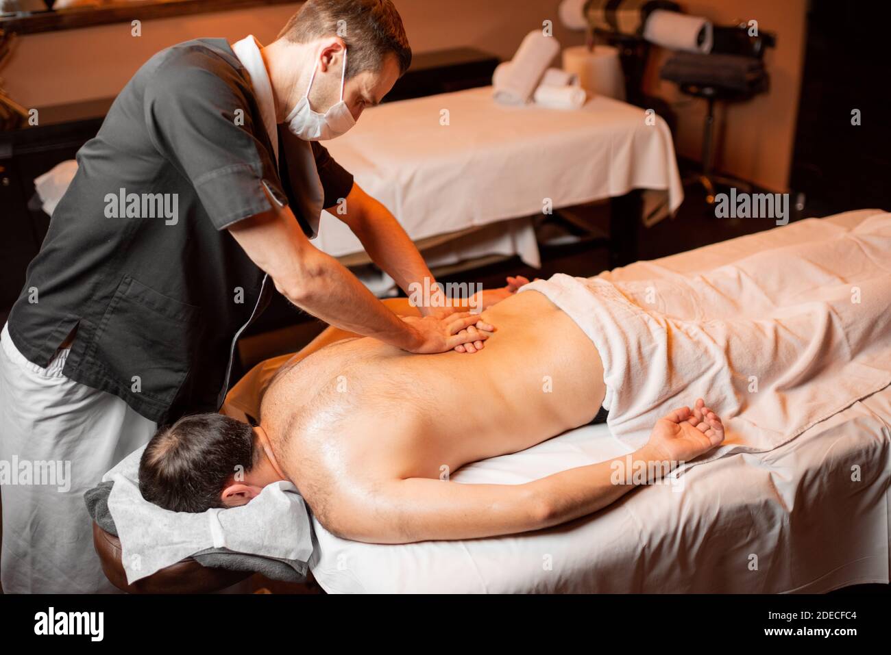 Massaggiatore professionista in maschera facciale che effettua un massaggio profondo ad un cliente maschile al salone Spa. Business durante il concetto epidemico Foto Stock