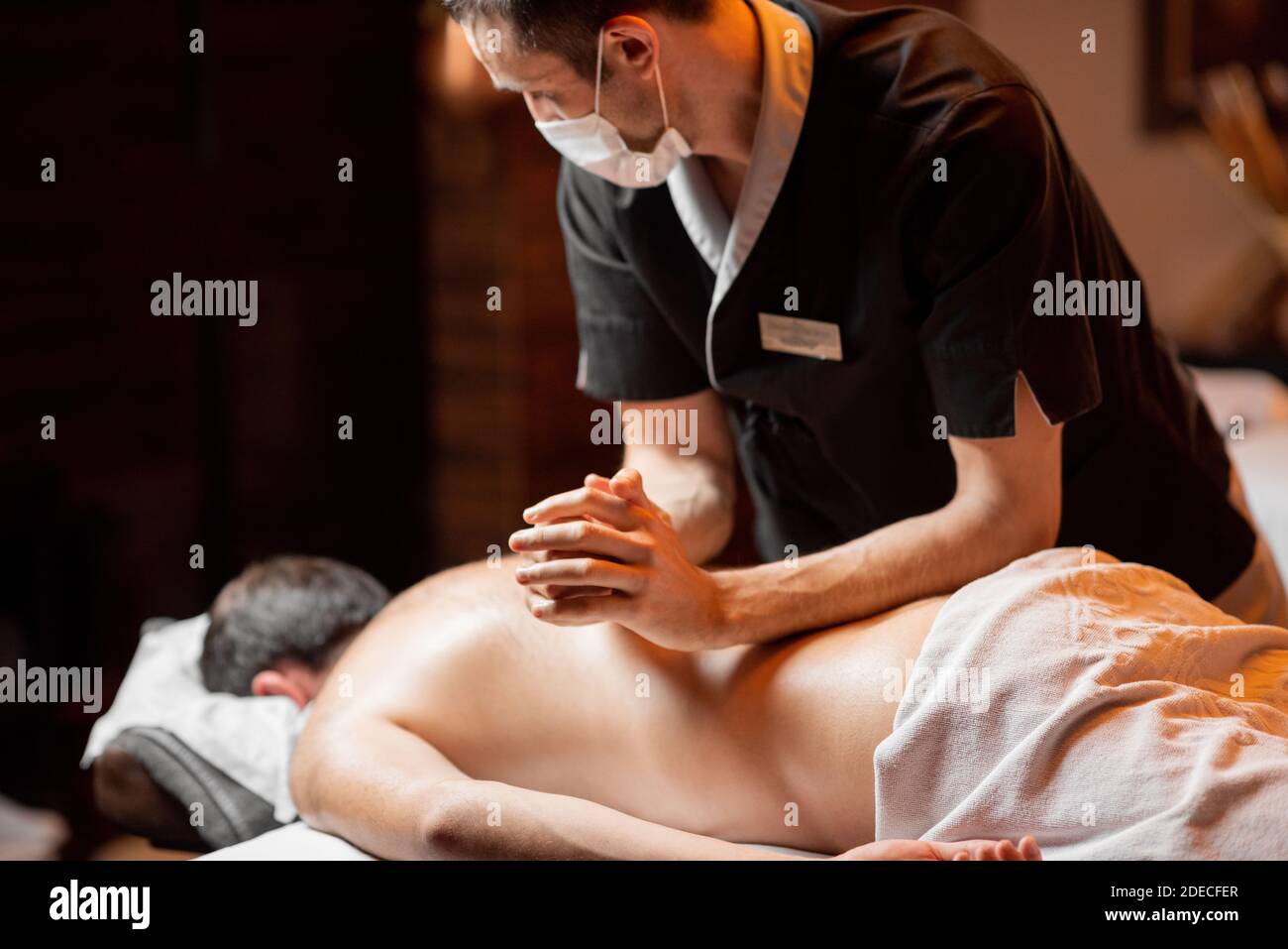 Massaggiatore professionista in maschera facciale che effettua un massaggio profondo ad un cliente maschile al salone Spa. Business durante il concetto epidemico Foto Stock